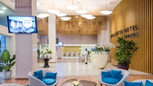 Eine Hotellobby mit einer Rezeption, einer Sitzecke mit Sesseln, einer großen Topfpflanze und einem Fernsehbildschirm, der eine Ankündigung zur Eröffnung einer Sky Bar zeigt.