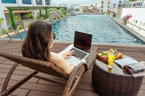 A person using a laptop on a wicker chair by a pool with a table of food and drinks.