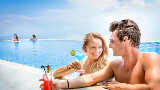 Ein Paar entspannt am Pool mit Getränken, im Hintergrund ist der klare Blick auf den Ozean zu sehen.