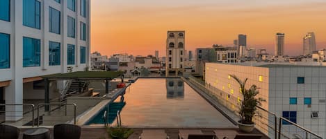 A rooftop pool with lounge chairs, a staircase, and a cityscape view at sunset.
