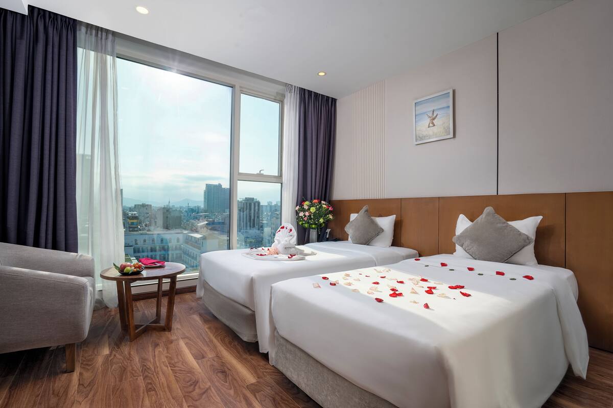 Habitación de hotel con dos camas, una mesita con un florero y vista a la ciudad a través de amplios ventanales.