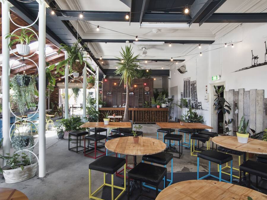 A cafe with round wooden tables and black stools, surrounded by potted plants and hanging planters.