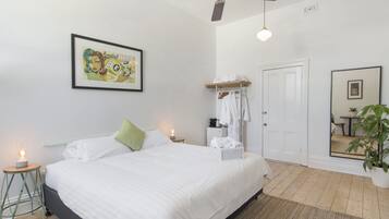 A bedroom with a bed, a ceiling fan, a bedside table with a lamp, a framed picture on the wall, a coat rack with clothes, and a potted plant.