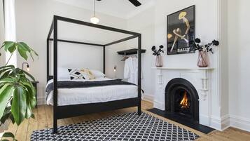 A four-poster bed with a black frame, a patterned rug, a fireplace with a white mantel, and a poster on the wall.