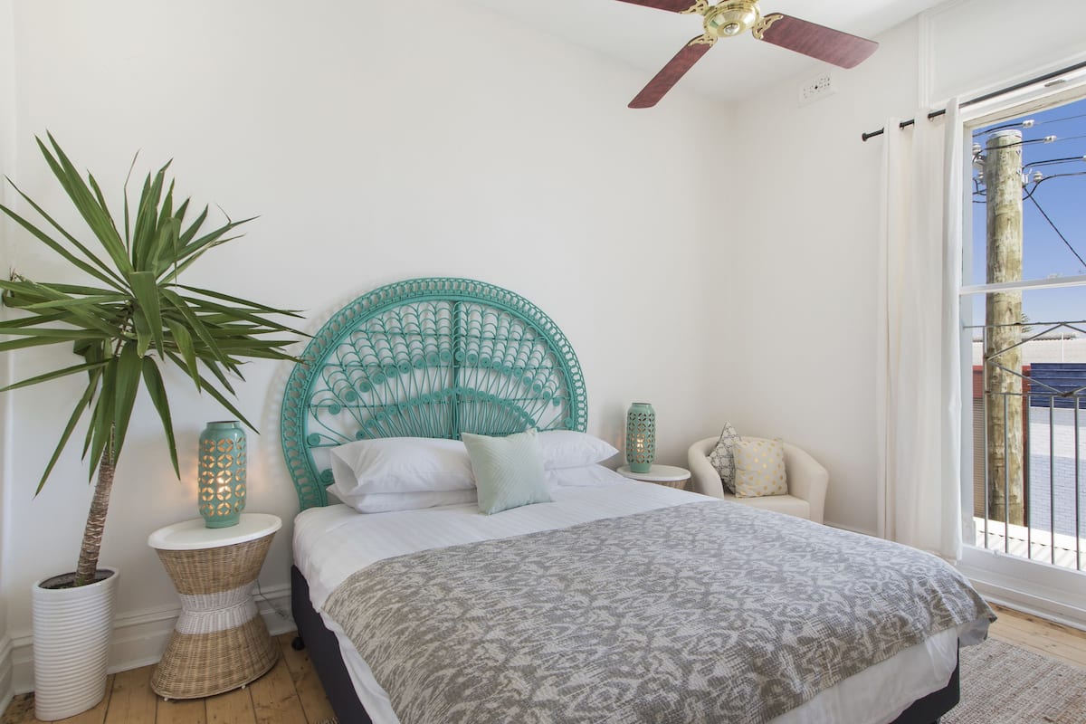 A bedroom with a bed, a large potted plant, a wicker side table, a decorative lamp, and a view of the outside through a window.