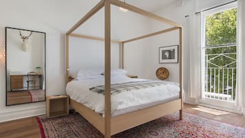 A four-poster bed with a wooden frame, a red patterned rug, a small bedside table, a mirror, a framed deer head, and a window with a view of trees.