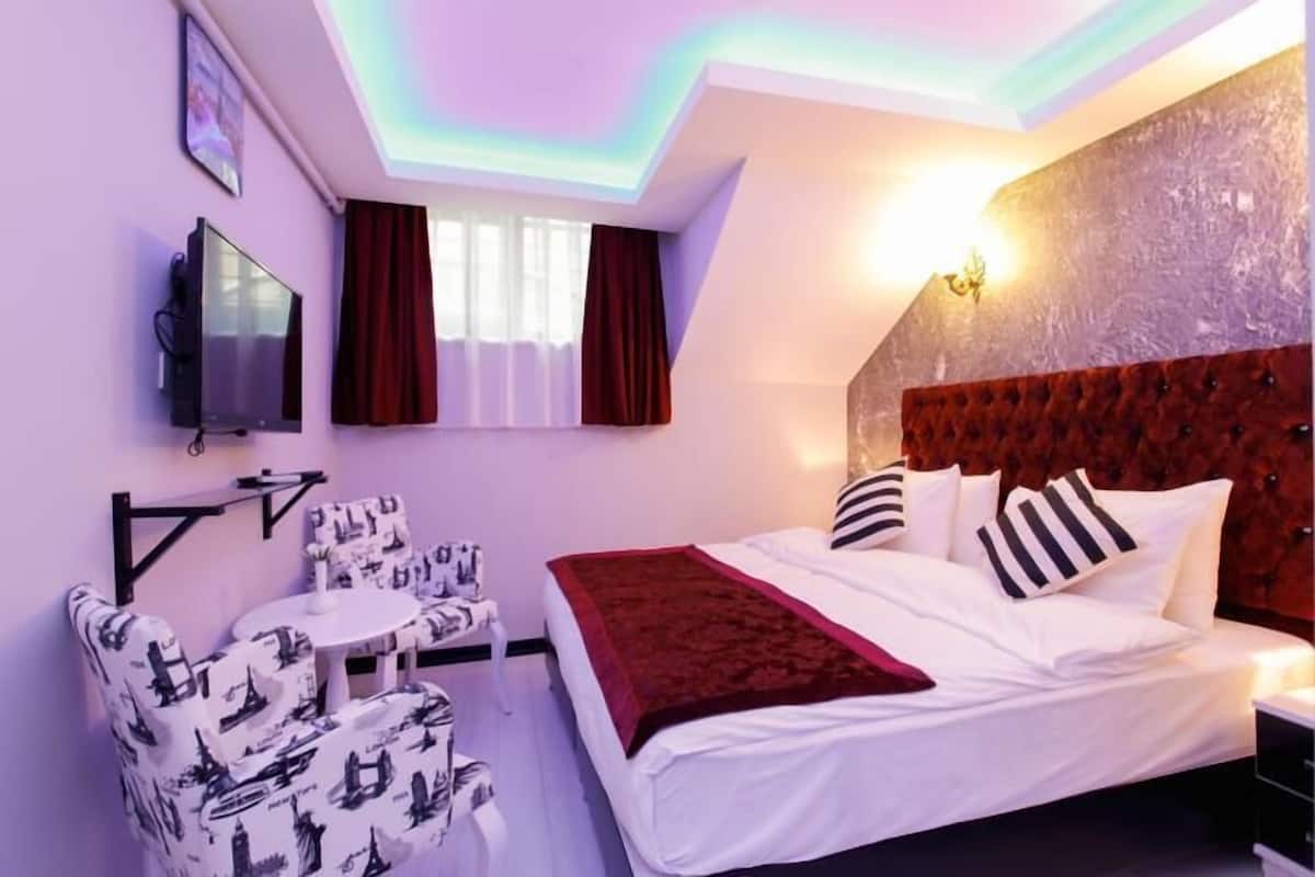 A hotel room with a bed, a TV, a small table with chairs, and a window with curtains.