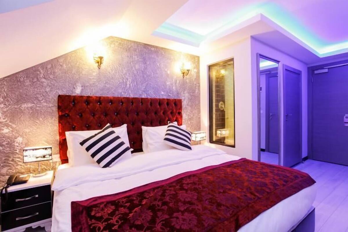 A modern hotel room with a tufted red headboard, white bedding, and a red patterned throw. There are two bedside lamps and a nightstand with a phone.