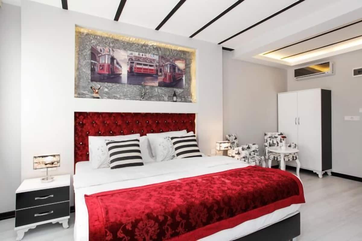 A modern bedroom with a red upholstered headboard, a large painting above the bed, and a white wardrobe.