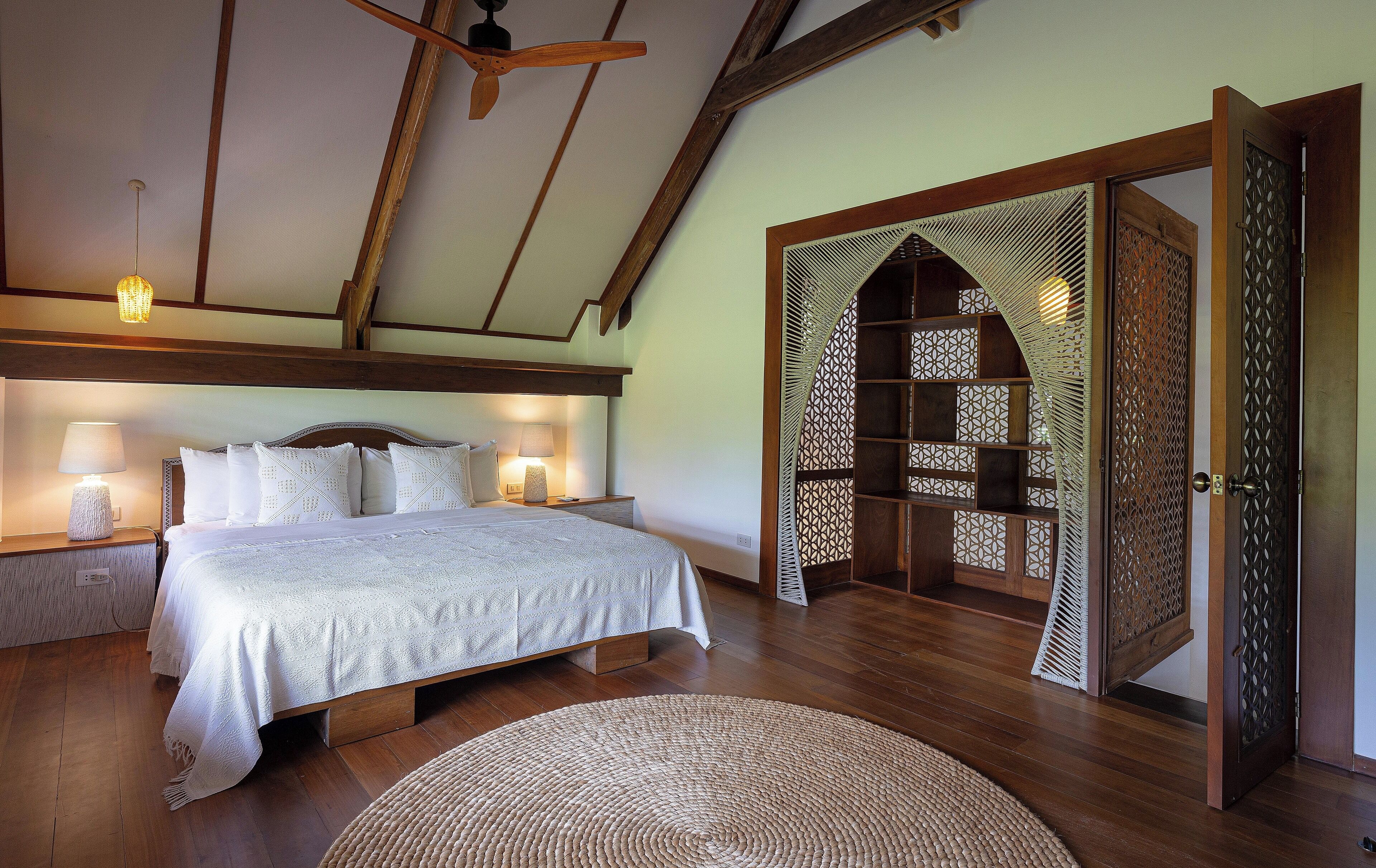 A bedroom with a wooden ceiling, a bed with white bedding, two bedside lamps, and a woven rug.