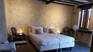 A bedroom with a stone wall, a bed with white bedding, two bedside tables with lamps, a wooden bench, and a standing fan. - Château Latour (Fours)