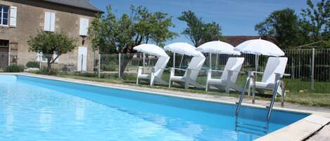 Une piscine entourée d’une clôture et d’un bâtiment doté de fenêtres et d’une cheminée, avec des chaises longues blanches et des parasols.