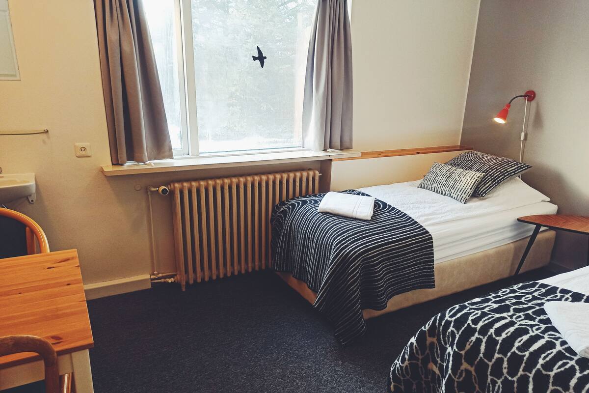 Une chambre d’hôtel comprenant un lit, un bureau, une chaise, une fenêtre avec des rideaux, un radiateur et une lampe de chevet.