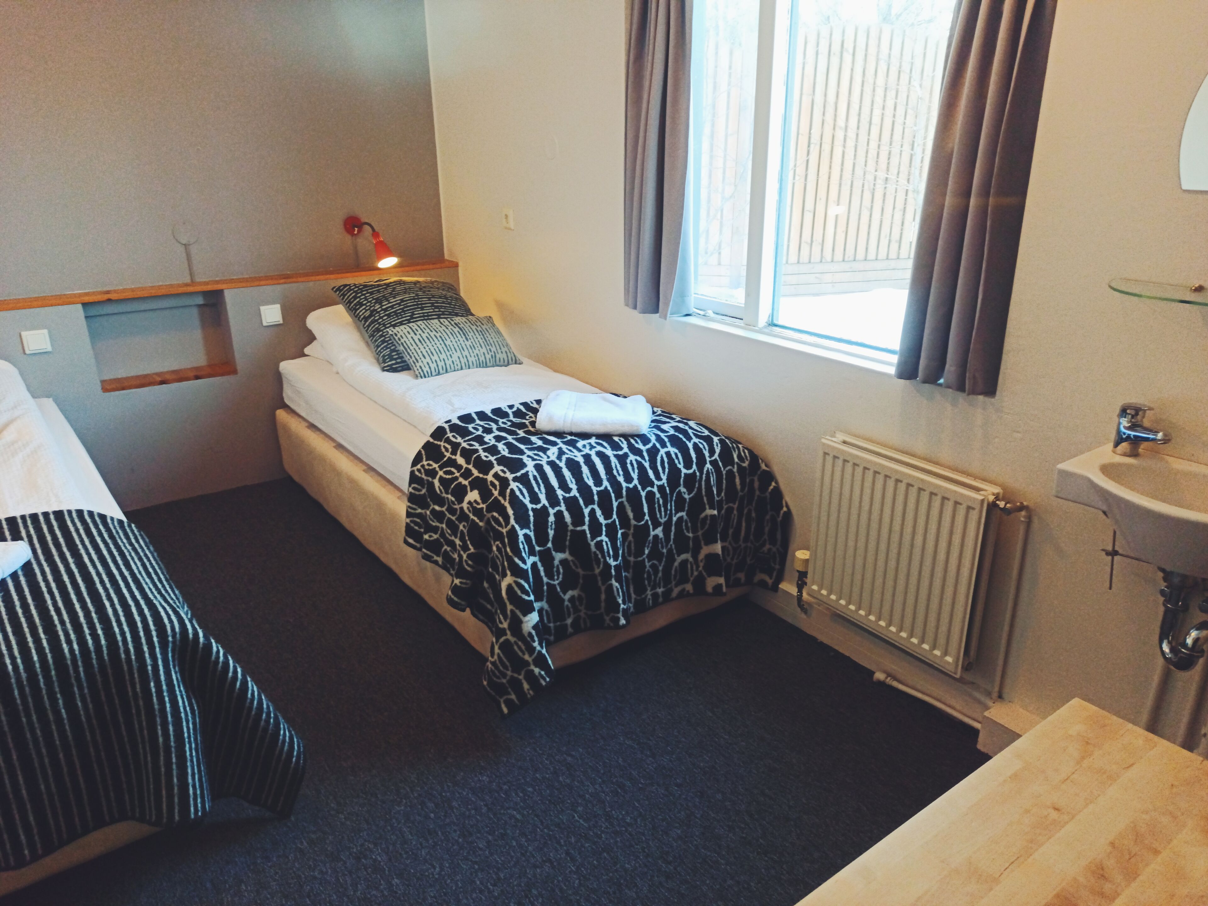 Une petite chambre d’hôtel avec un lit simple, une table de chevet, une lampe rouge, une fenêtre avec des rideaux, un radiateur et un lavabo.