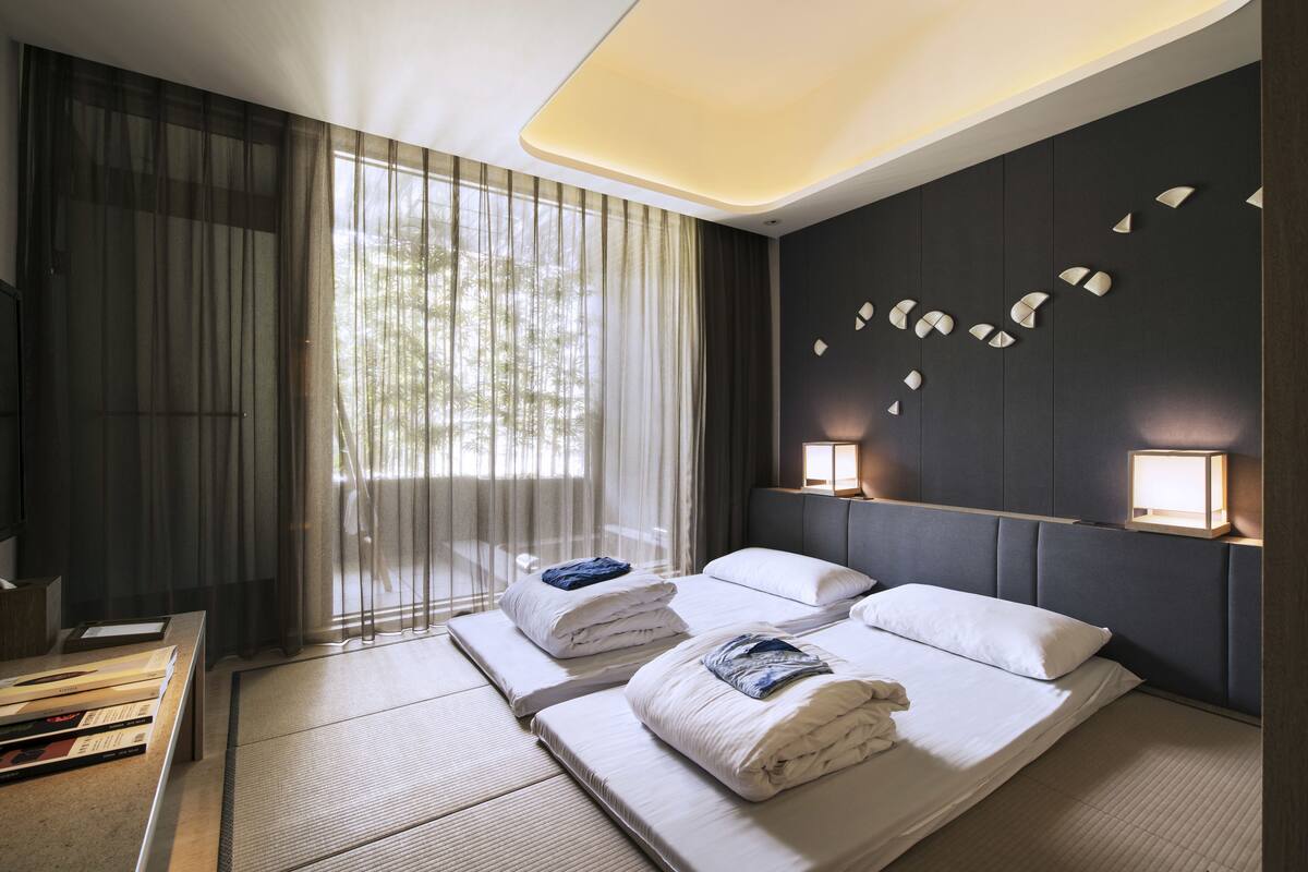 Ett modernt hotellrum med två sängar, ett stort fönster med tunna gardiner, ett nattduksbord med böcker och en väggmonterad lampa.