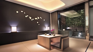 Een moderne hotelkamer met een houten eettafel, een bank en een groot raam met uitzicht op bamboeplanten.