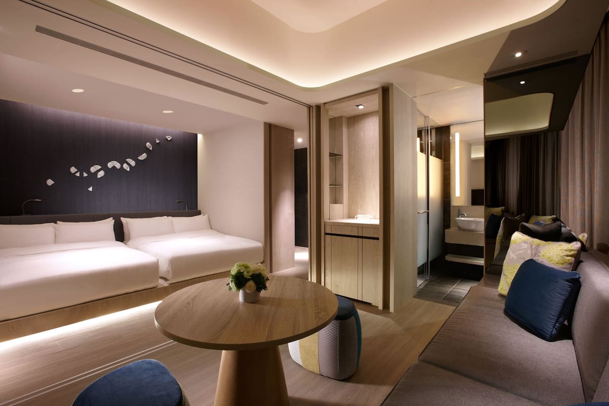 Ett modernt hotellrum med en soffa, ett runt bord med en växt, en liten köksdel och ett badrum med spegel och handfat.