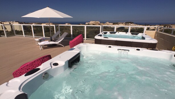 Un jacuzzi en una terraza con sillones de descanso y una sombrilla, con vistas a un paisaje urbano costero.