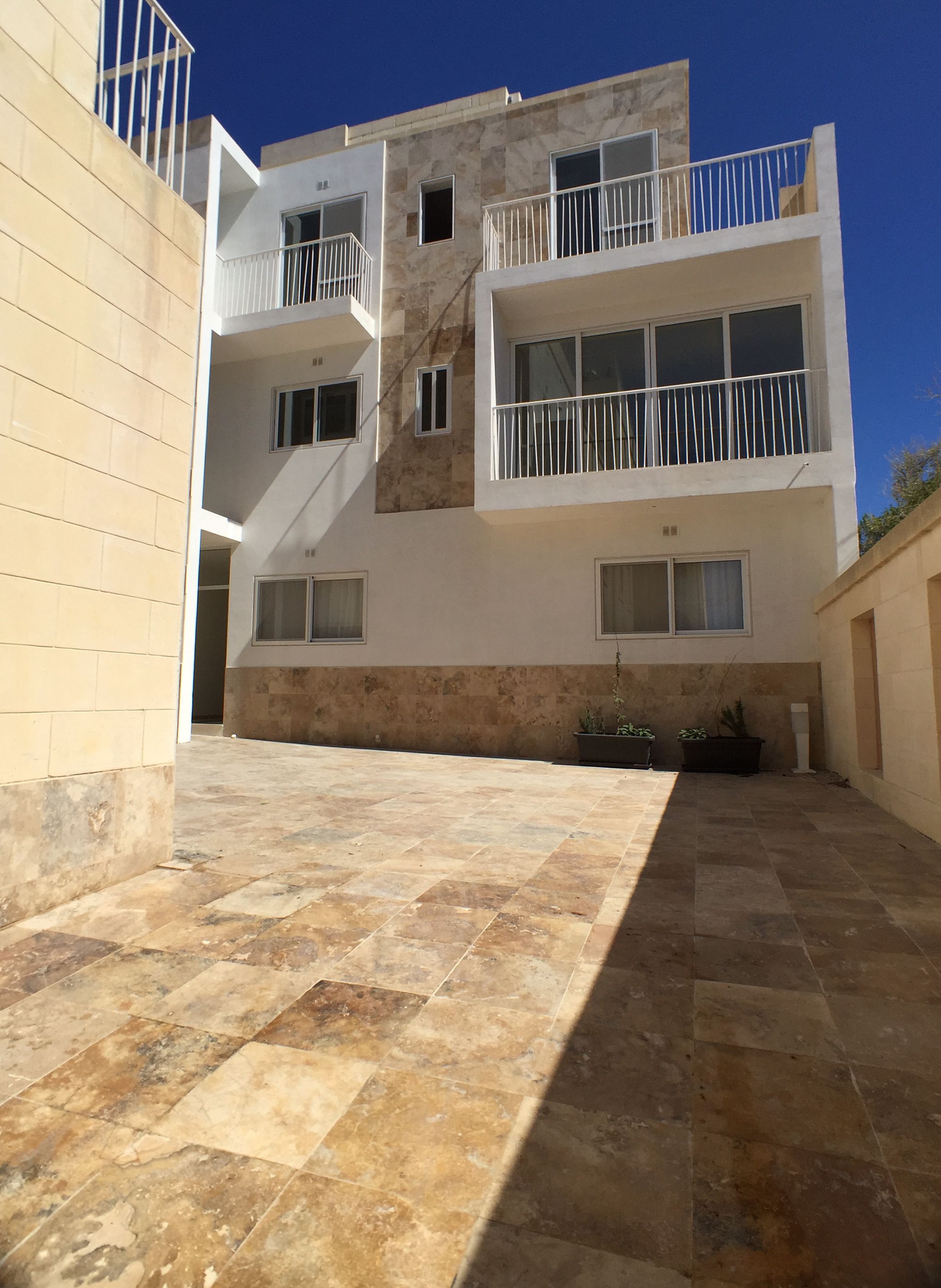 A modern building with a courtyard, featuring a mix of beige and white walls, multiple balconies with white railings, and a tiled floor.