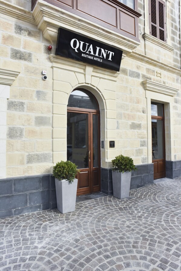 A building with a sign that reads "QUAINT BOUTIQUE HOTELS" above the entrance. There are two potted plants on the ground in front of the entrance. - Quaint Boutique Hotel Xewkija (Xewkija)