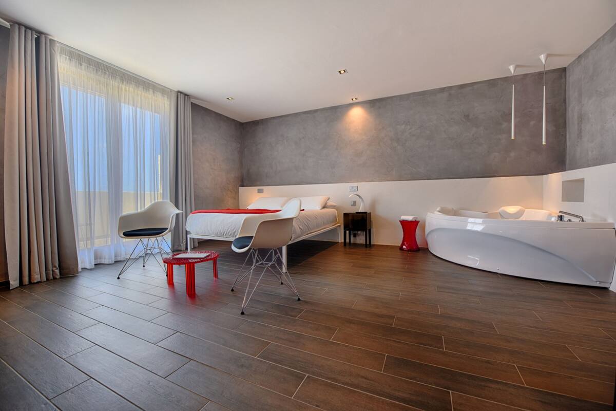 A modern hotel room with a large bed, a sitting area with chairs, a small table, a red stool, a bathtub, and a large window with curtains.
