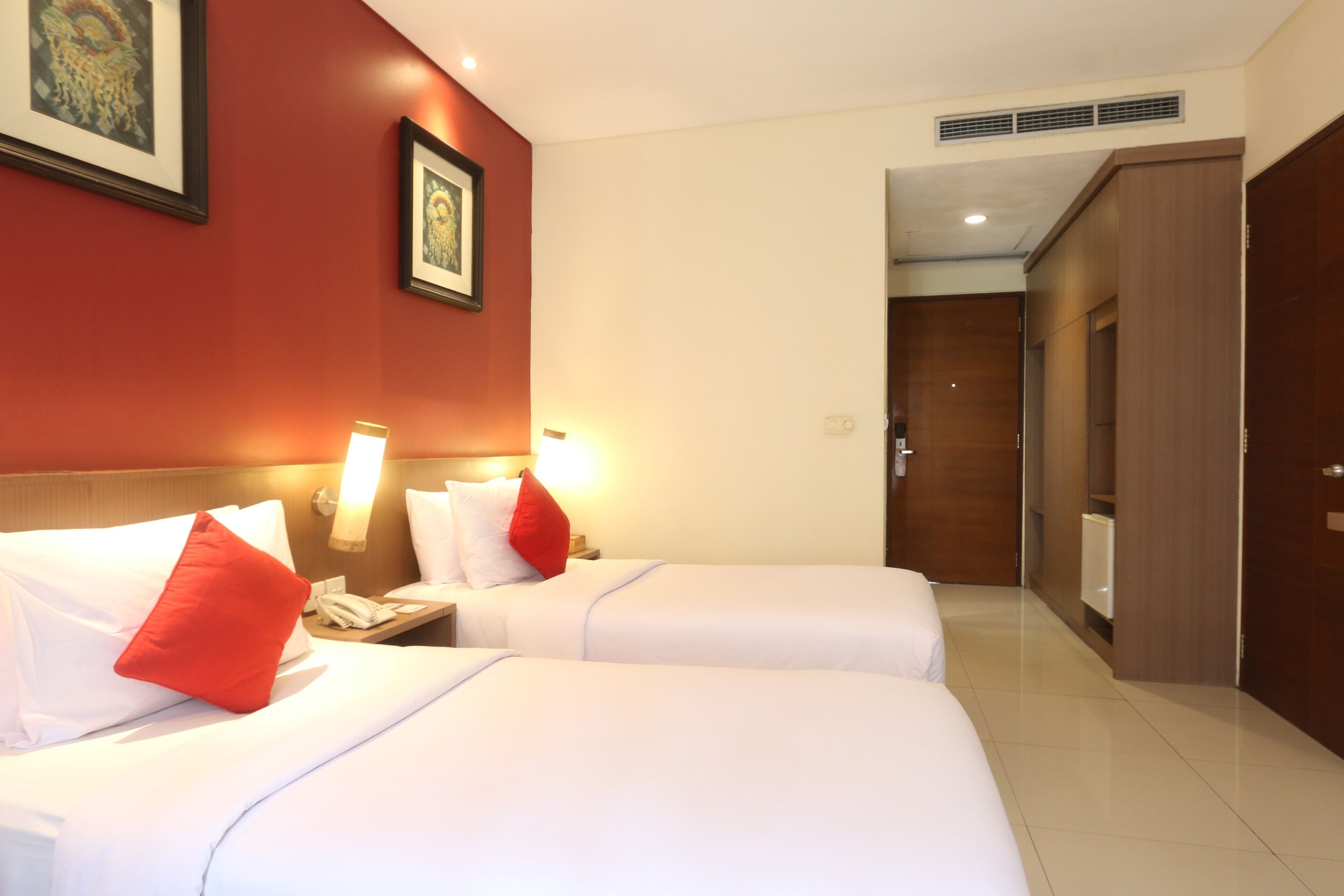 Habitación de hotel con dos camas, almohadas rojas, una mesita de noche con teléfono y obras de arte enmarcadas en la pared.