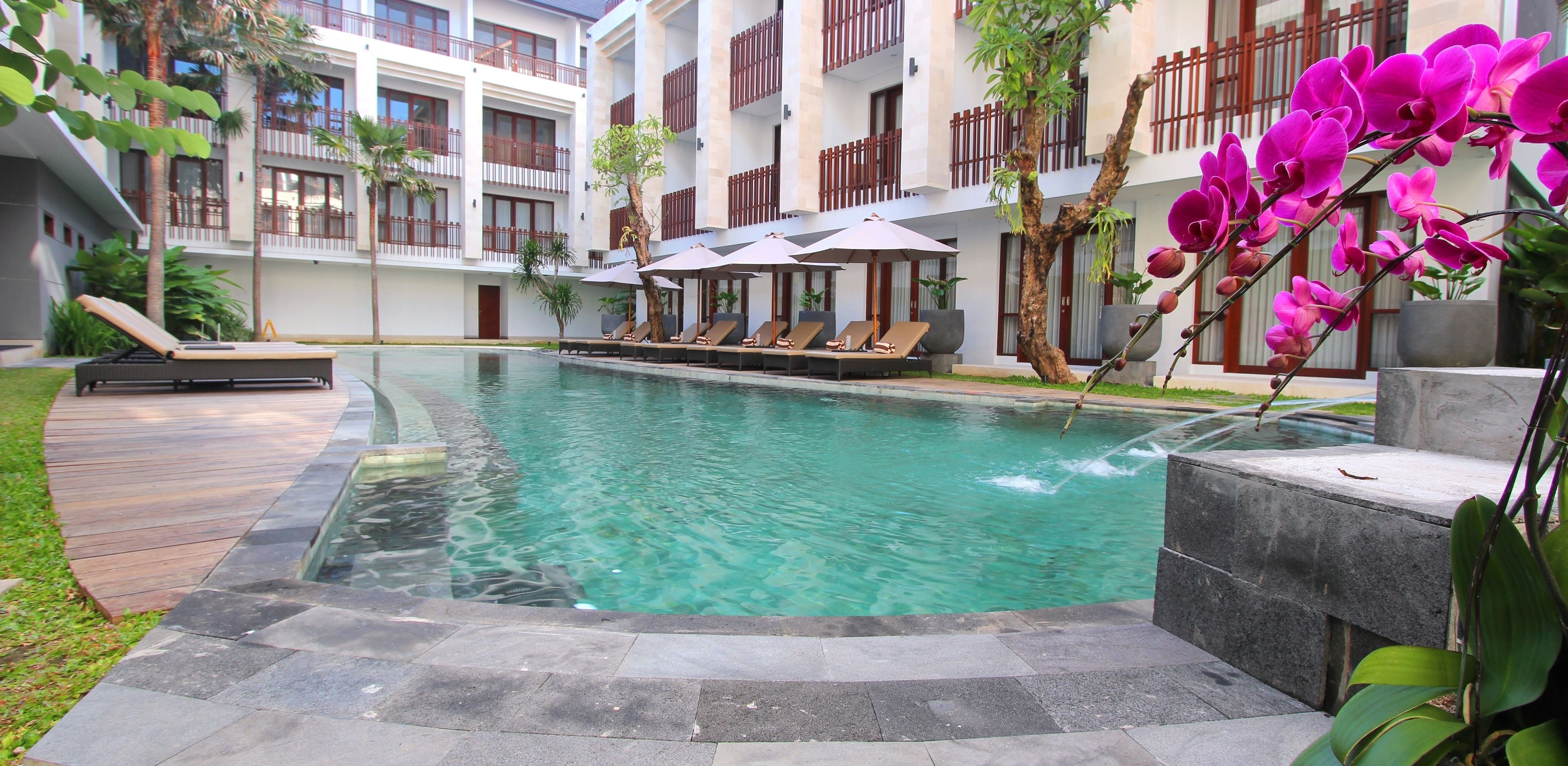 A hotel pool area with lounge chairs and umbrellas, surrounded by greenery and a building with balconies.