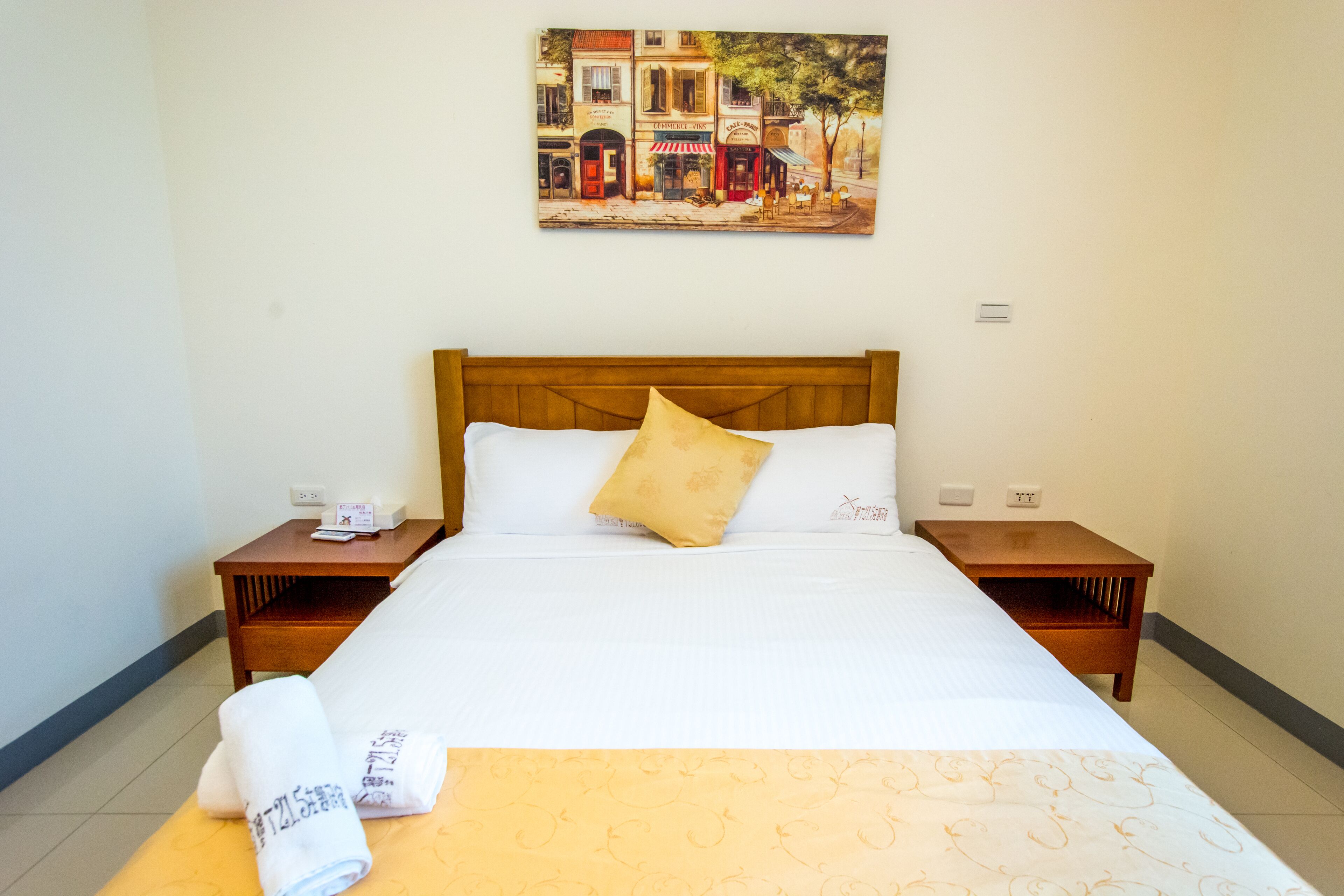 A hotel room with a large bed, a wooden headboard, two nightstands, a painting on the wall, and a neatly made bed with a yellow pillow.