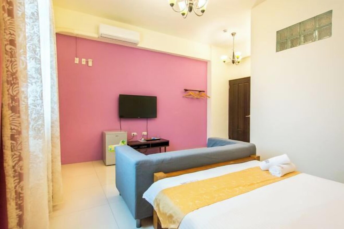A hotel room with a pink accent wall, a flat-screen TV, a bed with a yellow blanket, a blue armchair, and a small desk with a lamp.