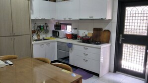 A compact kitchen with white cabinets, a microwave, a stove, and a sink. There is a dining table with chairs and a purple mat. - Kenting MuChen INN (Hengchun)