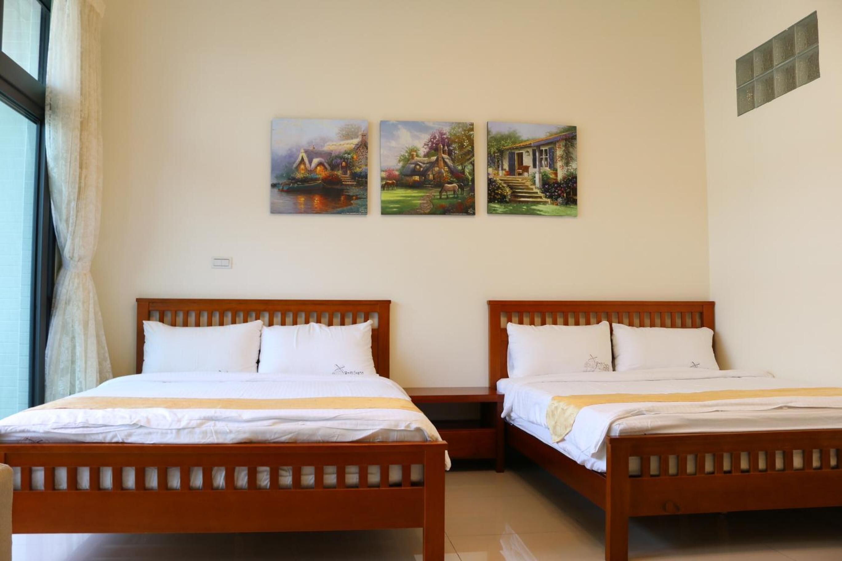 A hotel room with two wooden beds, a window with curtains, and three paintings on the wall.
