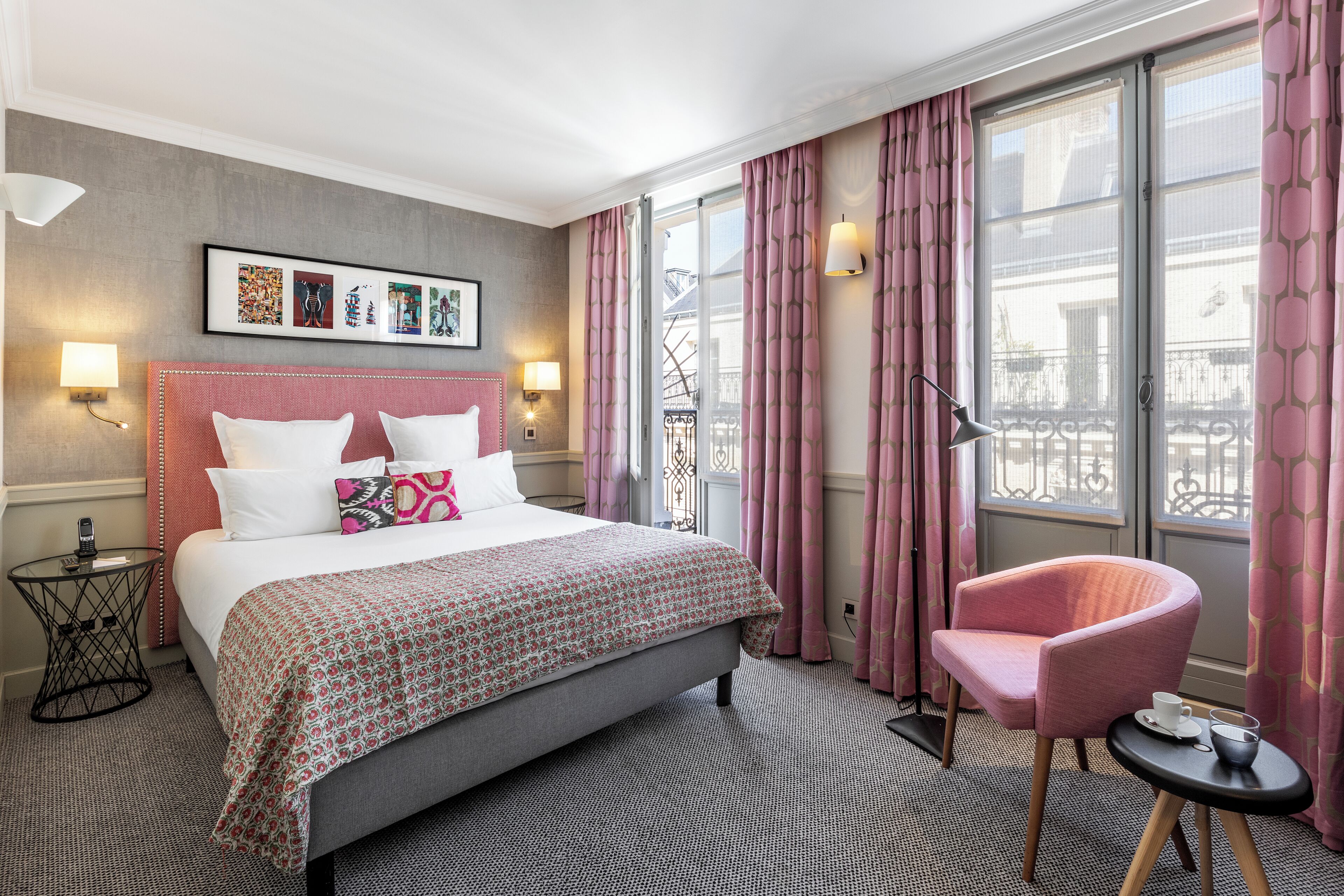 Een hotelkamer met een groot bed, een roze fauteuil, een klein tafeltje met een kopje en een raam met gordijnen.
