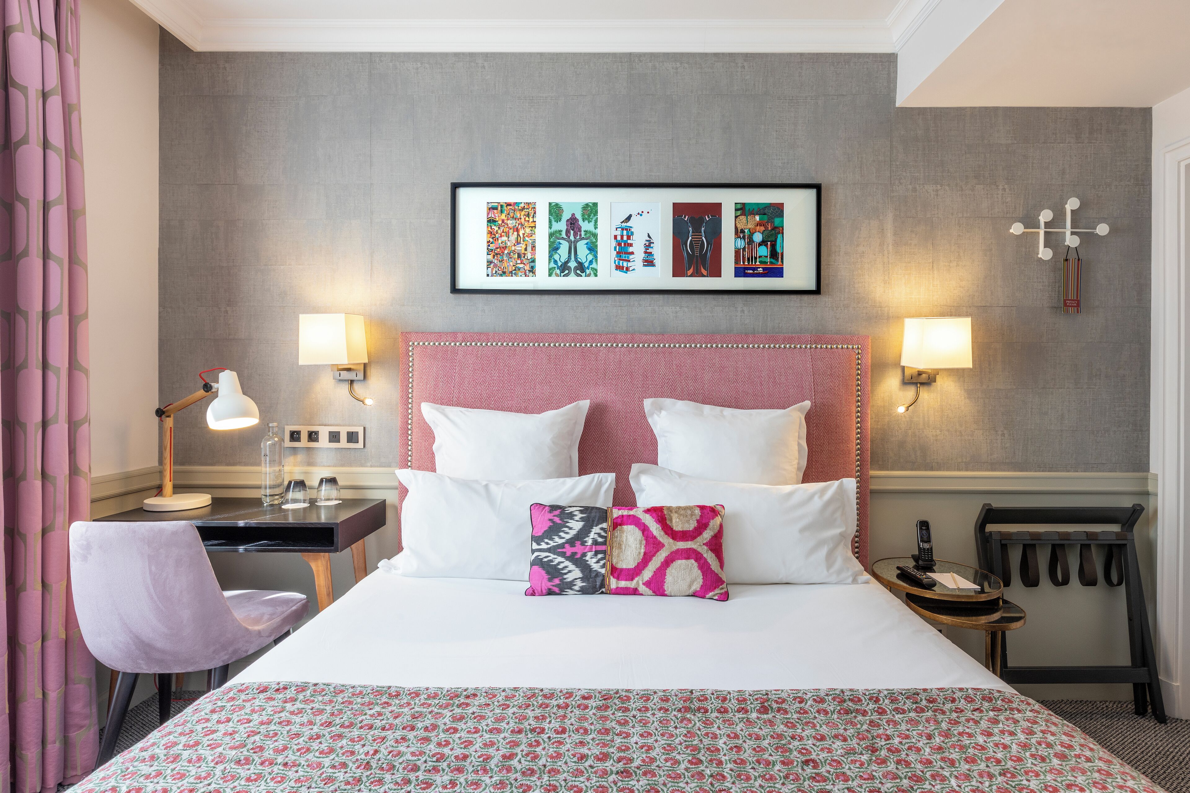 A hotel room with a bed, two bedside lamps, a desk with a lamp, a chair, a framed artwork on the wall, and a coat rack.
