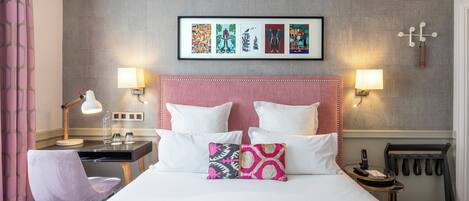 A hotel room with a bed, two bedside lamps, a desk with a lamp, a chair, a framed artwork on the wall, and a coat rack.