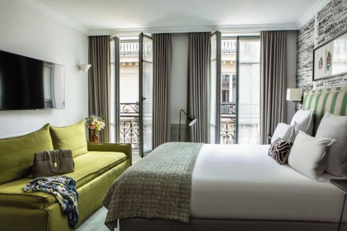 Una habitación de hotel con cama, sofá verde, televisor y balcón con cortinas.