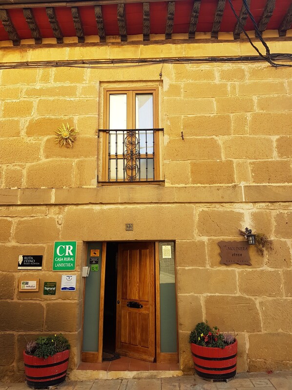 A rustic building with a wooden door, a window with decorative iron bars, and two flower pots on the ground.