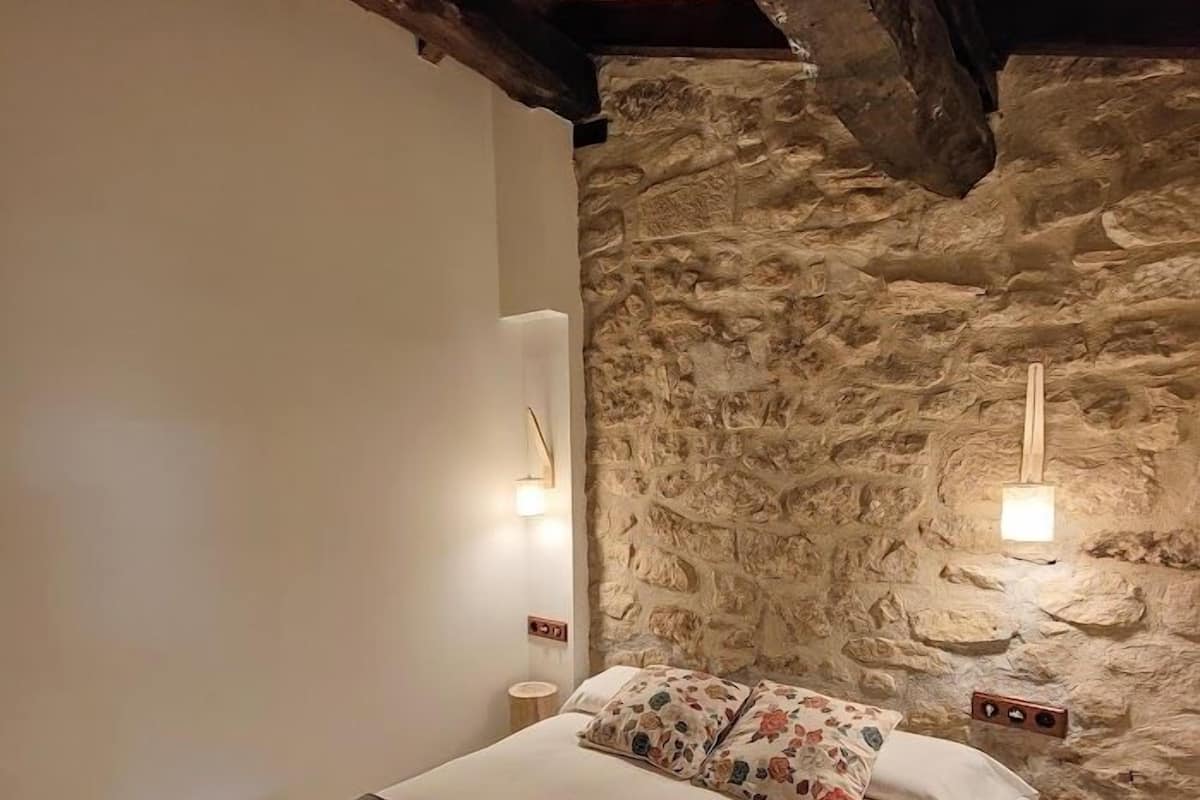 A bedroom with a stone wall, wooden beams, a bed with floral pillows, and two wall-mounted lights.