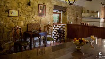 A rustic stone wall with a framed certificate, wooden chairs, a table with fruit and glasses, and a bar area with shelves.