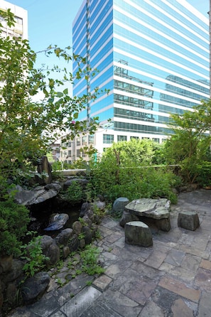 Un sendero de piedra con un pequeño elemento acuático y rocas, rodeado de follaje, que conduce a un edificio moderno con ventanas de vidrio reflectante.