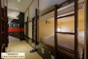 Habitación de hotel con literas, piso de madera, letrero de salida montado en la pared y una cama con edredón blanco.