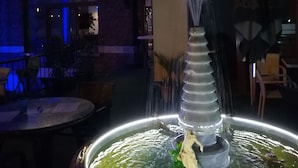 An outdoor fountain with a statue in the center, surrounded by a circular pool. In the background, there is a building with a sign that reads "Flyinn Hotel & Lounge." - Fly Inn Brussels Airport (Machelen)