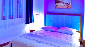 A bedroom with a bed, a nightstand, a window with curtains, a wall-mounted picture, and a ceiling light. - Fly Inn Brussels Airport (Machelen)