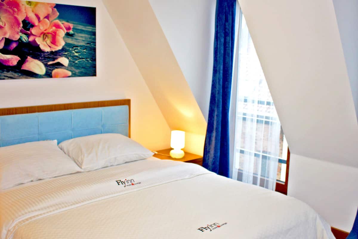 A neatly made bed with a headboard, a bedside lamp, and a wall-mounted painting of flowers.