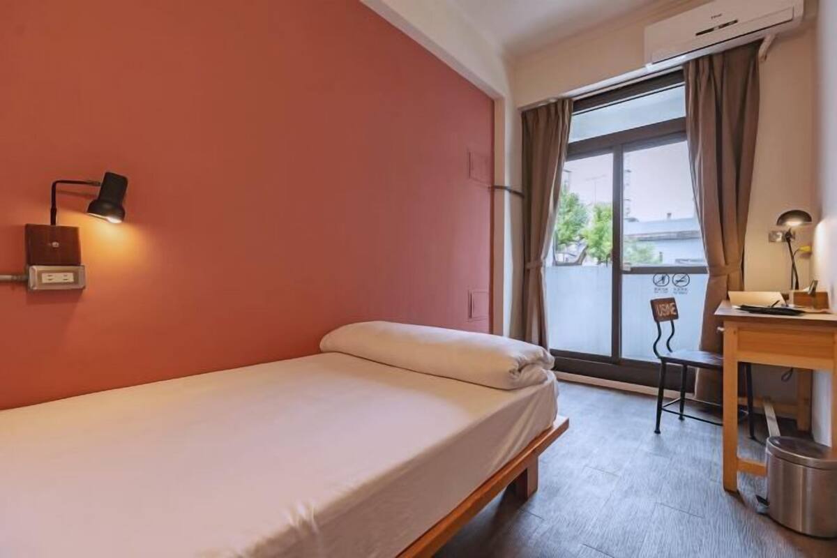 Een eenpersoonsbed met hoofdeinde, een bureau met stoel, een lamp en een airconditioning.