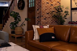 A living room with a brown leather sofa, a potted plant, and a brick wall.