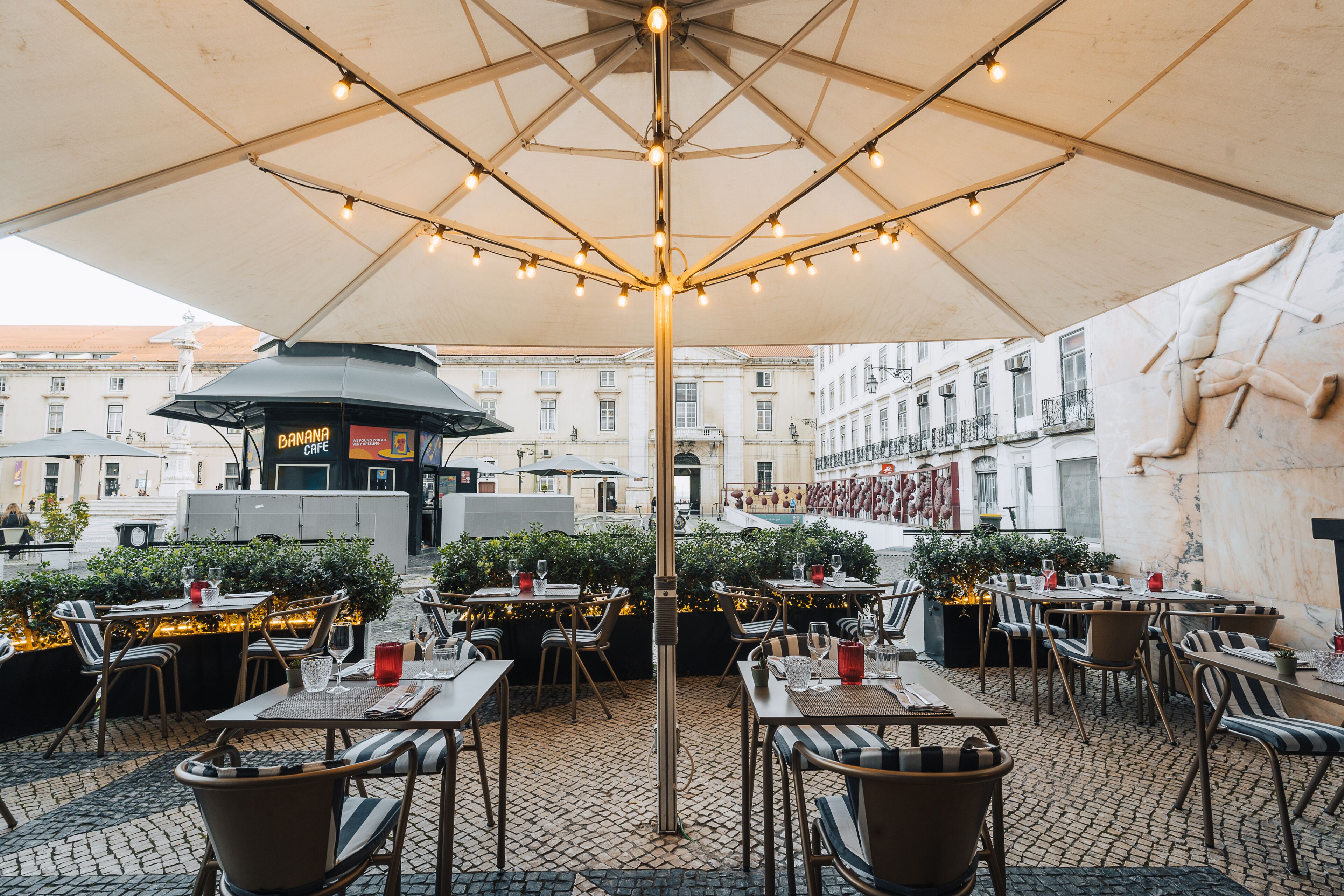 An outdoor café with tables and chairs, a large umbrella, and a building in the background.