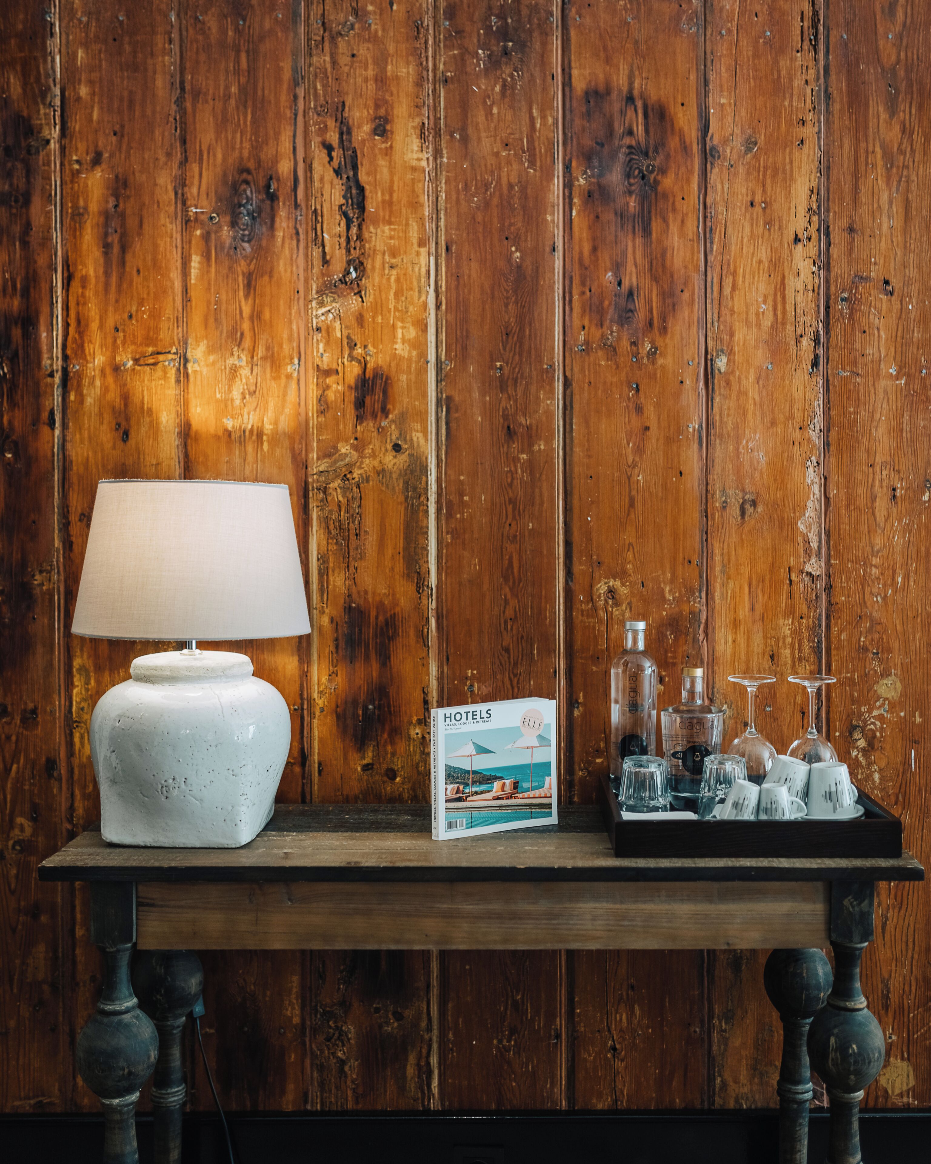 A wooden table with a white lamp, a magazine, and a tray with glasses and bottles.