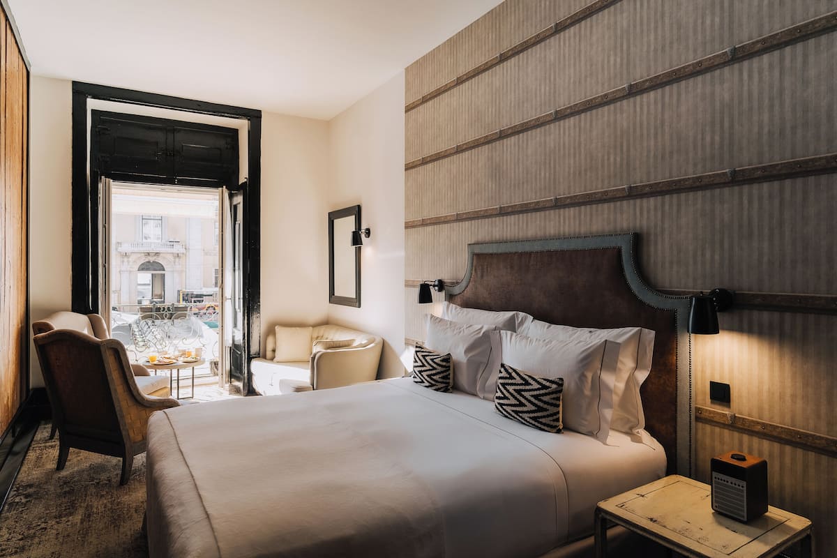 A hotel room with a bed, a chair, a small table, a lamp, and a view of the outside.