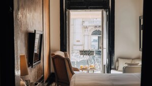 Habitación de hotel con cama, una silla, una pequeña mesa con desayuno, televisor y balcón con vista a edificios.