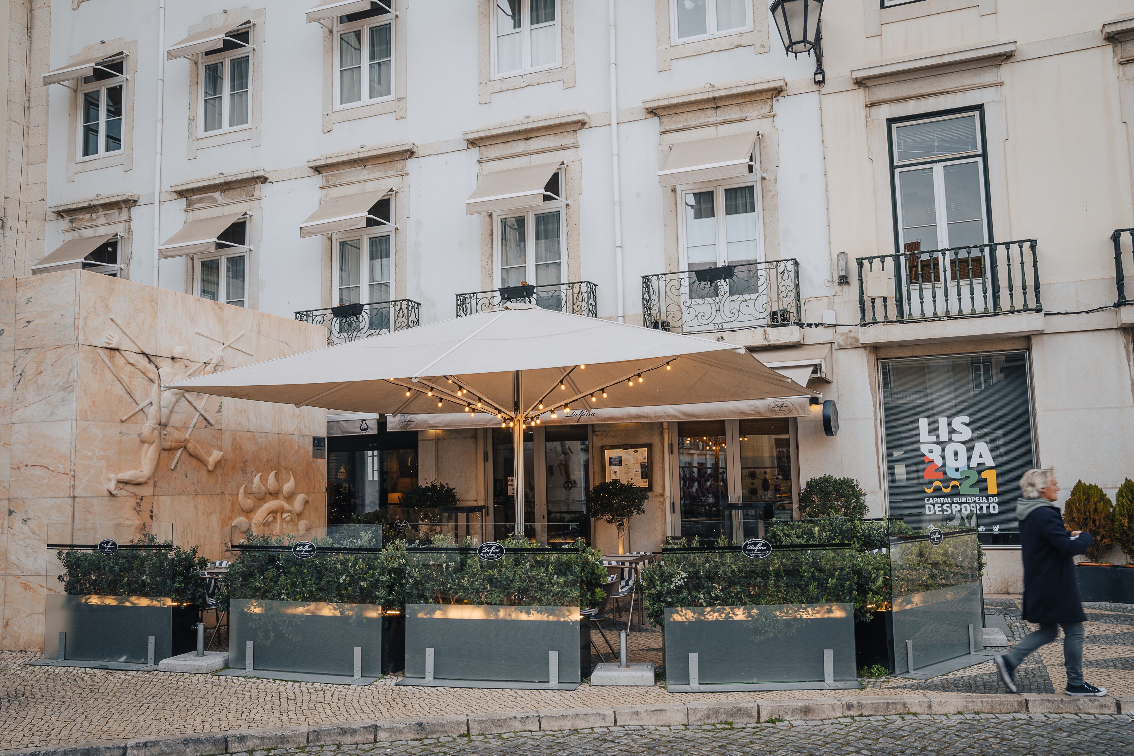 Un bâtiment doté d’une grande terrasse, une enseigne indiquant « Lisboa 21 », et une personne qui passe.